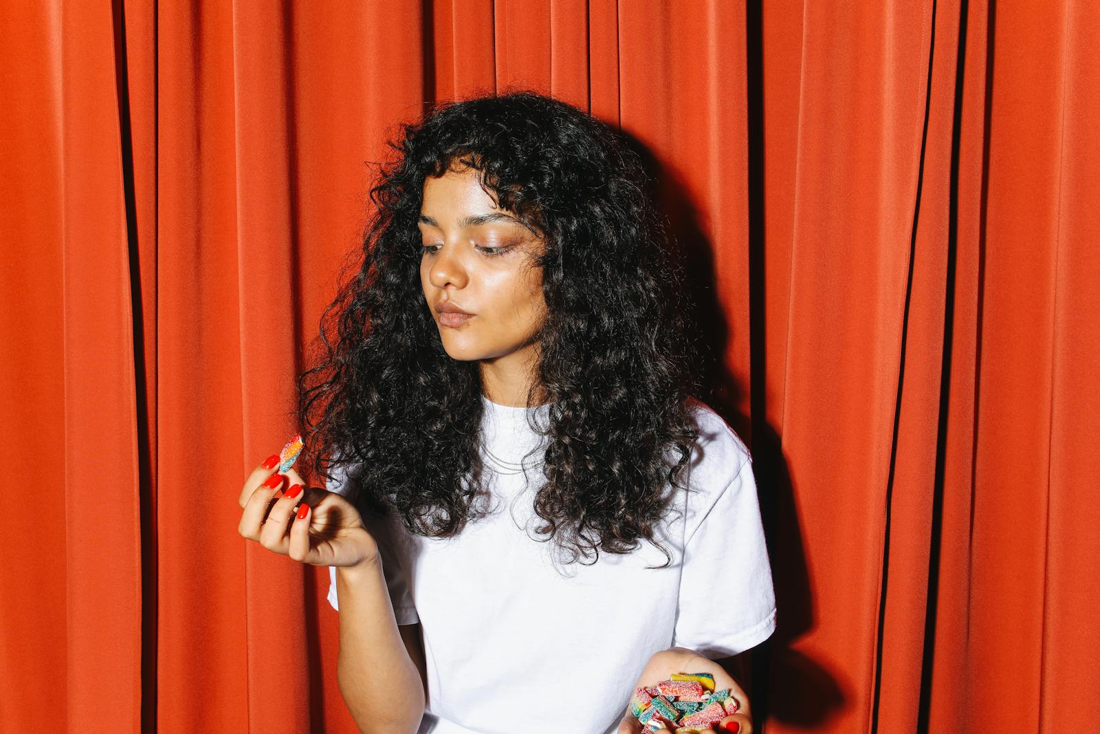 A woman with curly hair holding colorful gummy candies with a red curtain backdrop.