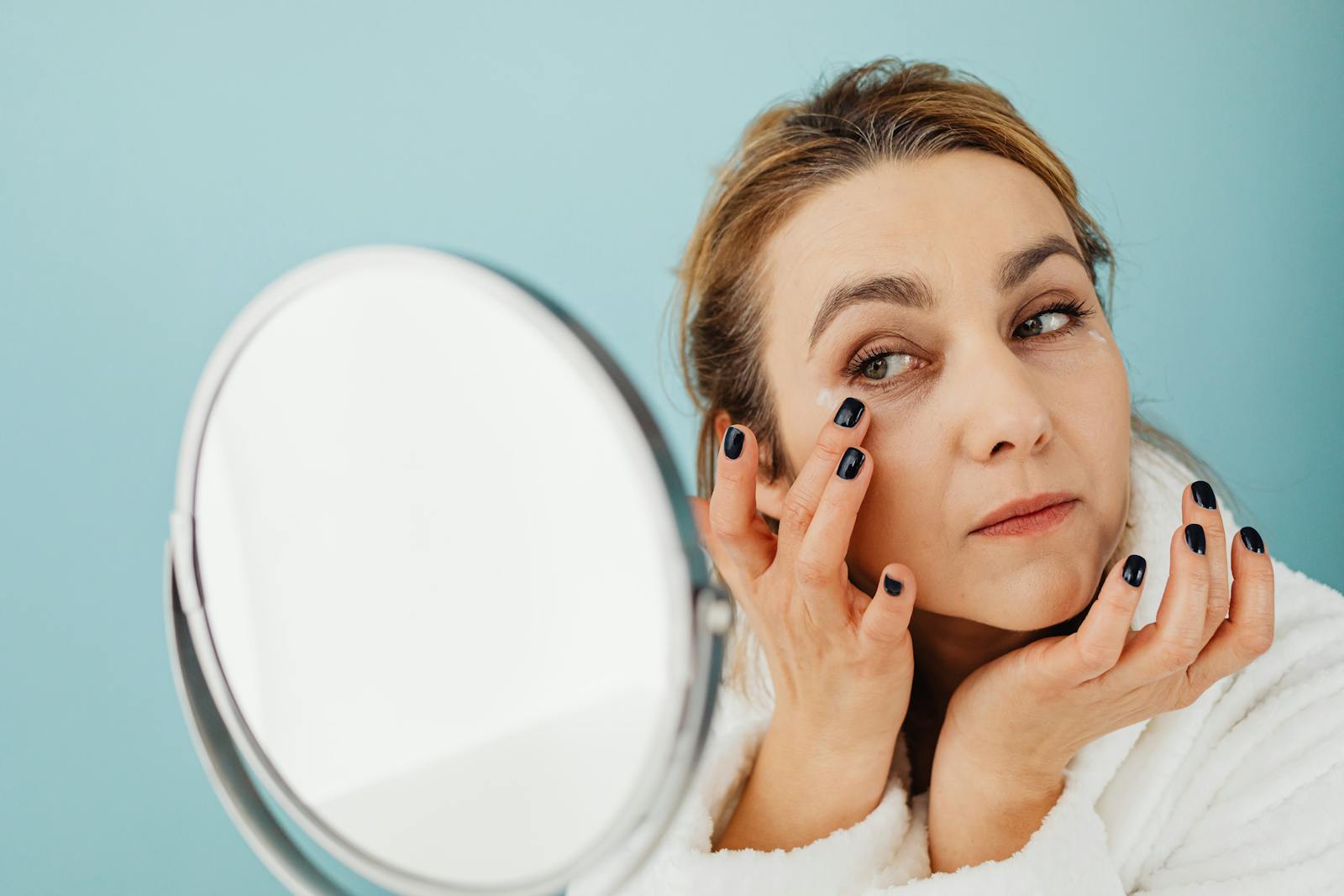 A woman in a bathrobe applies skincare cream while looking in the mirror. Ideal for beauty and self-care concepts.